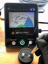 Load image into Gallery viewer, A close-up of a black SmartPro Gem-Eye1 gemstone simulator device on a wooden surface, with a gloved hand using a jeweler's tool to place or adjust a clear gemstone in a small black holder below the screen.

